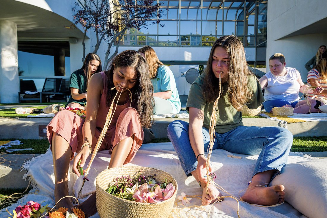 Ellice and another young women with an arm limb difference figure out a creative way to adaptively make their haku leis by holding one side of the string with their feet, the other side with their teeth, and weaving the flowers in with their one hand.