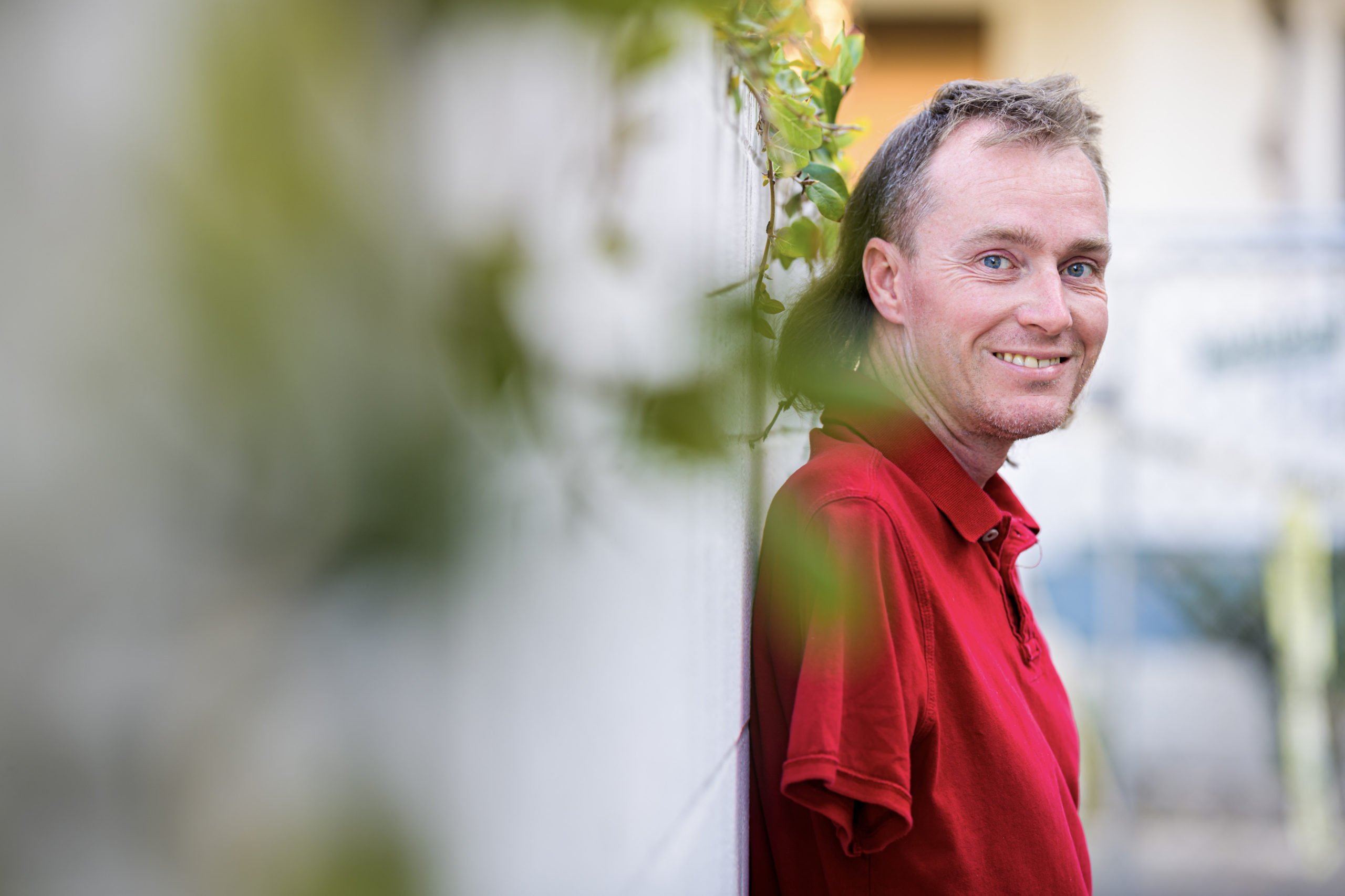 A portrait of Forge attendee Shane smiling while leaning against a wall with vines and showing off his right arm limb difference.