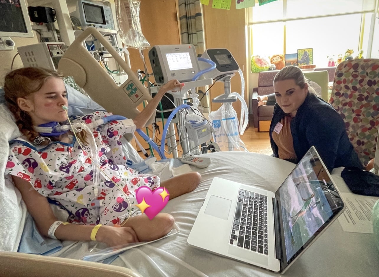 Ainsely sitting in her hospital bed watching one of the Beautifully Flawed retreat recap videos on a MacBook with Sarah Hill.