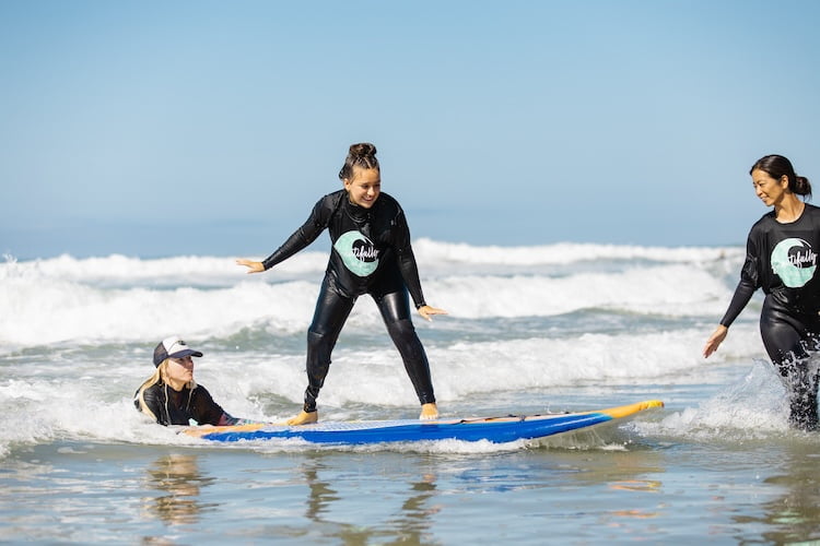 Beautifully Flawed attendee and right leg amputee Lily smiles as she stands up on her surfboard and rides a wave to shore.
