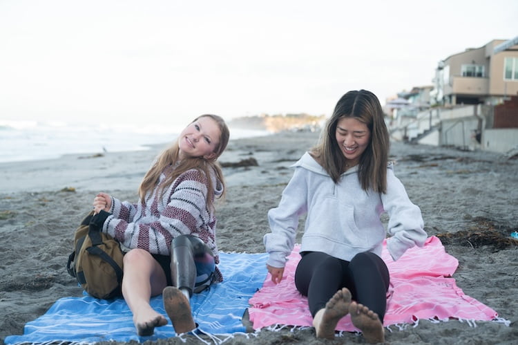 Two young women attendees attempt a partner workout on the beach where they pass a heavy sandbag back and forth to one another.