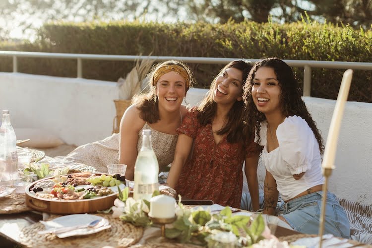 Three Beautifully Flawed attendees smile for a group photo while enjoying a beautiful bohemian picnic in a scenic backyard.
