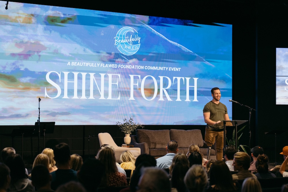 A man stands at a podium addressing the audience during the Shine Forth event with a large event screen behind him.