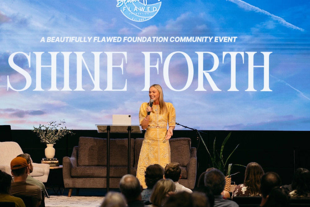 A woman in a yellow dress smiles while speaking on stage at the Shine Forth community event.