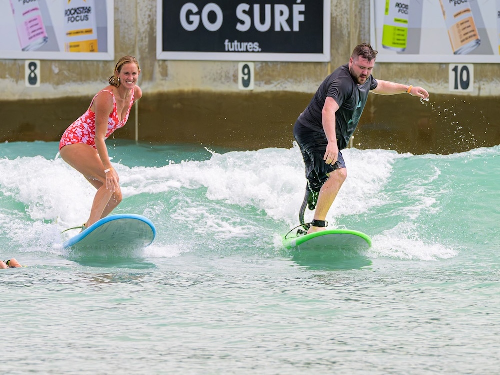 A man and woman, both with prosthetic legs, joyfully surfing side-by-side on a wave during a Beautifully Flawed Foundation activity